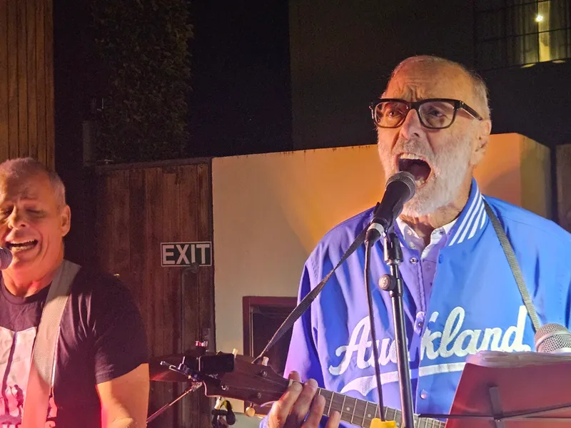 Two older men perform on stage; one sings passionately into a microphone while playing guitar and wears a blue "Auckland" jacket, the other smiles and sings. A wooden wall and "EXIT" sign are in the background.