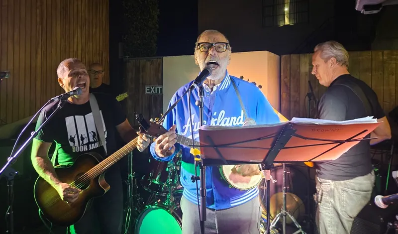 A band performs on stage at night; a man in an Auckland jacket sings and plays tambourine, others play guitar, bass, and drums, with sheet music on a stand and an EXIT sign visible in the background.
