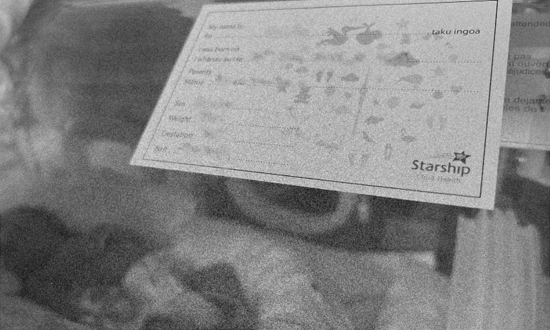 A black and white photo showing a newborn baby lying in a hospital bassinet with a Starship Child Health info card above, displaying blank spaces for name, birth date, weight, height, and other details.