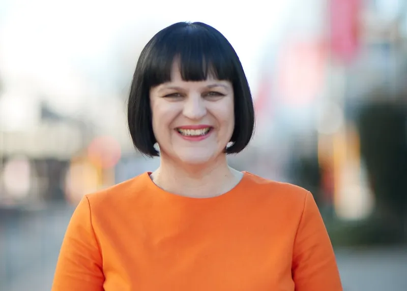 a woman with a dark brown bob haircut smiles at the camera wearing a bright orange dress