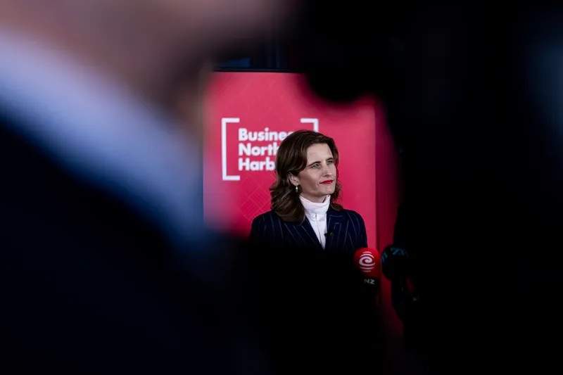 A woman in a dark suit stands in front of a red backdrop with the words “Business North Harbour.” She appears to be speaking at an event, with blurred figures and microphones in the foreground.