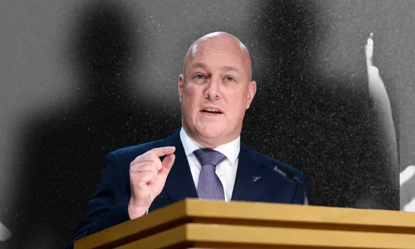 A bald man in a suit and tie speaks at a podium, gesturing with his hand. The background is black and white with blurred shadows of people.