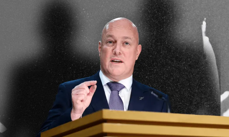 A bald man in a suit and tie speaks at a podium, gesturing with his hand. The background is black and white with blurred shadows of people.