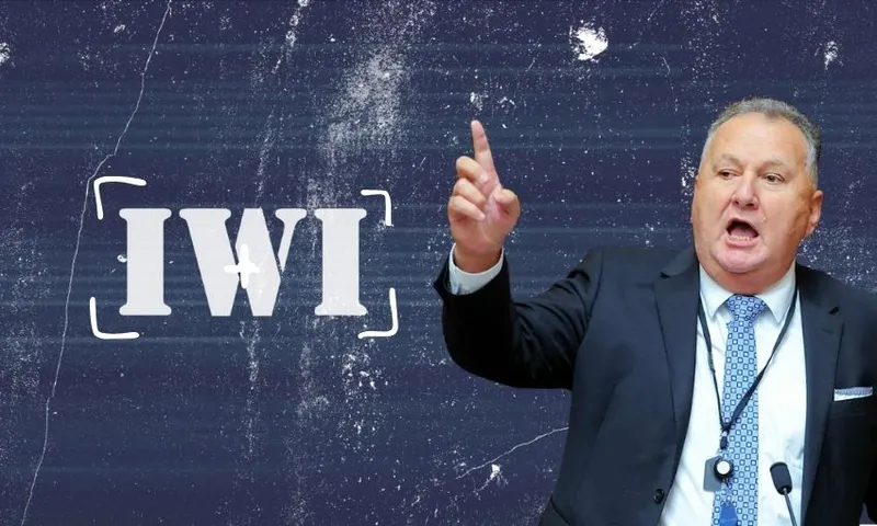 A man in a suit gestures with his right hand while speaking in front of a textured dark background with the word "IWI" in bold white letters beside him.