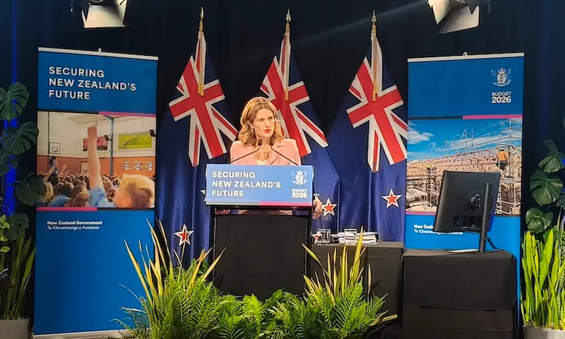 A woman stands at a podium labeled "Securing New Zealand's Future" with New Zealand flags behind her, flanked by two banners and a monitor, speaking at what appears to be a government event or press conference.