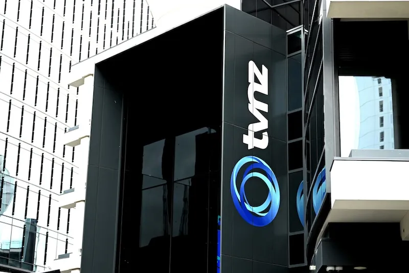 The photo shows the exterior of a modern building with the TVNZ logo and name displayed on a black wall. Surrounding buildings are reflected in the glass and visible in the background.