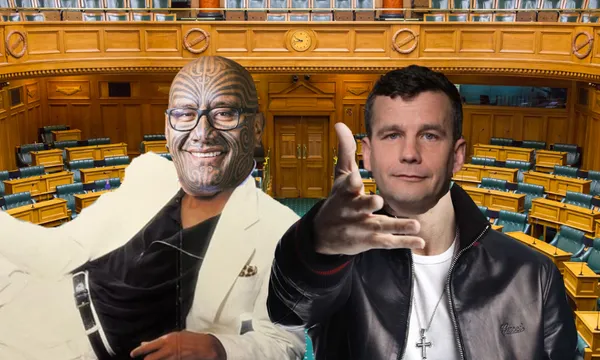 Two men pose in front of empty wooden parliamentary chamber seats; one wears a white suit and glasses with facial tattoos, the other wears a black jacket and gestures with his hand.