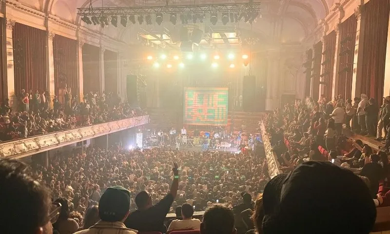 A large crowd fills a grand, ornate concert hall with balconies, watching a brightly lit stage where musicians perform. Warm lights and a colorful screen create a lively atmosphere.