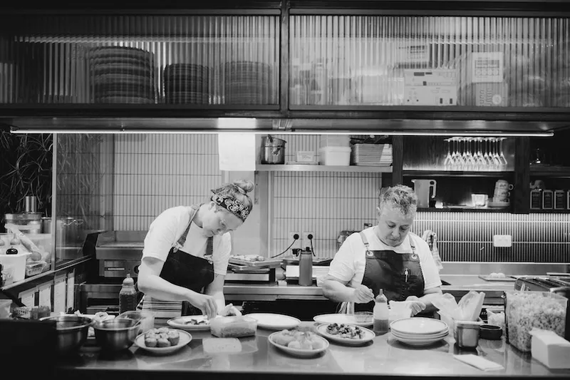 Two chefs in aprons prepare dishes in a restaurant kitchen. Various ingredients and plates are on the counter. Shelves with plates, glasses, and supplies are visible in the background. The scene is in black and white.