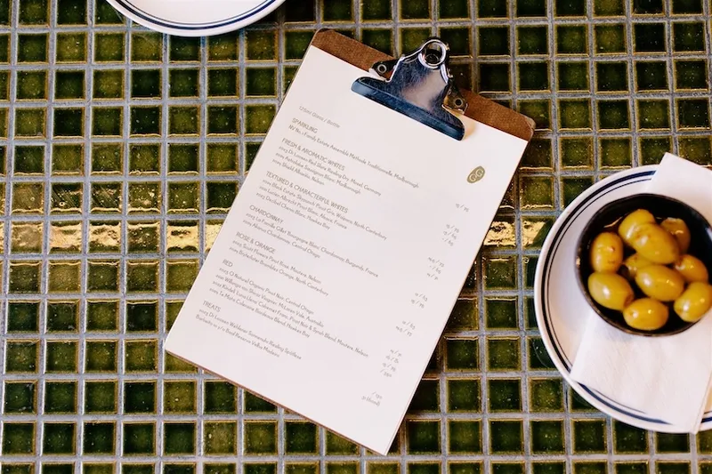 A restaurant menu on a clipboard is placed on a green tiled table next to a small bowl of green olives and a plate with a napkin.
