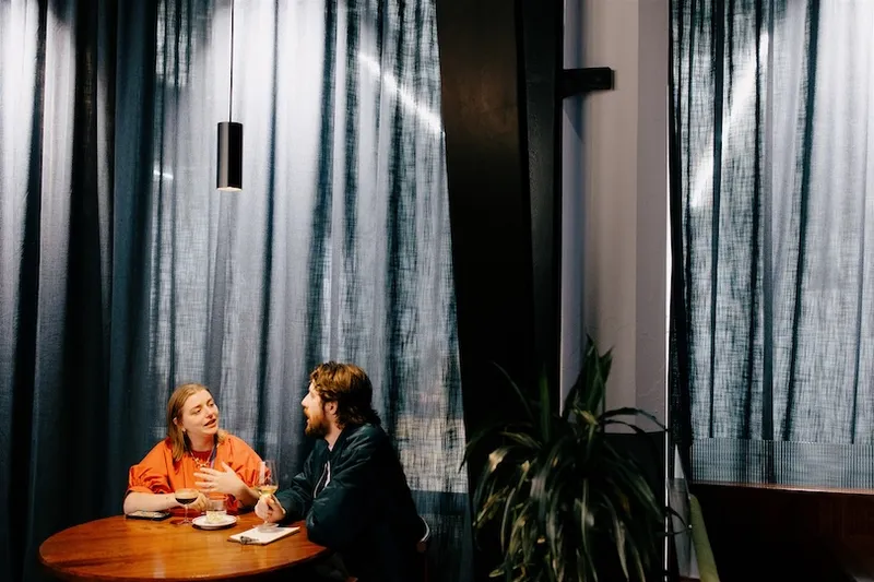 Two people sit at a round wooden table by a window with dark curtains, having a conversation. One is eating, and there is a plant in the foreground. A hanging light fixture is above the table.