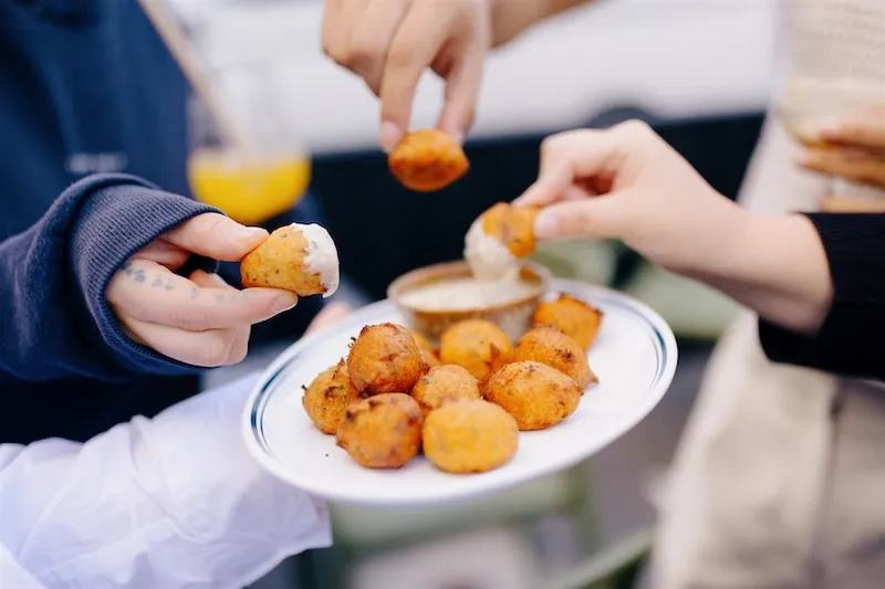 Three people reach for fried snacks from a white plate, dipping them into a sauce. One hand has tattoos and wears a blue sleeve. The scene appears to be at an outdoor gathering.