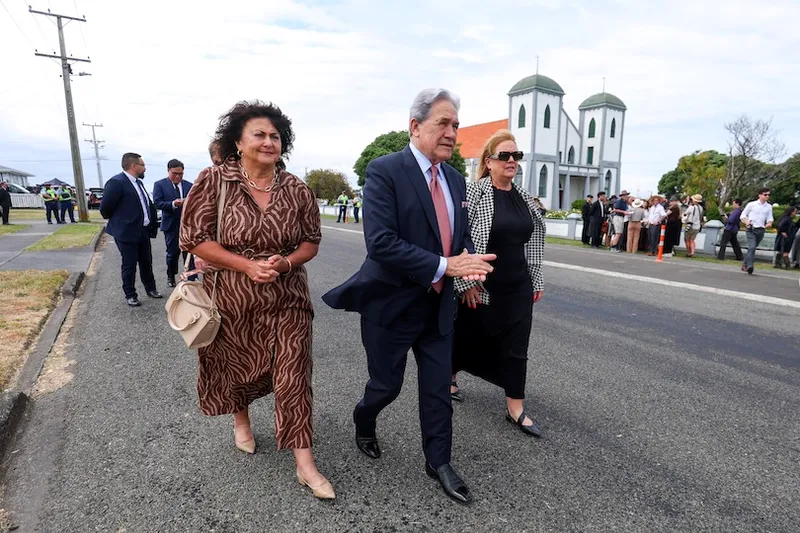 Three people walk together on a street, dressed formally, with a crowd and a church in the background. The man in the center wears a suit, while the two women on either side wear patterned dresses.