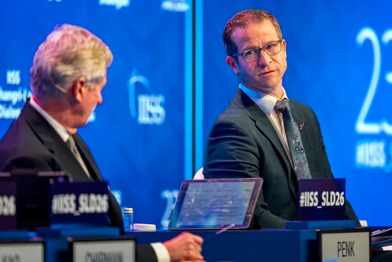 Two men in suits sit on a panel stage with blue backgrounds displaying “IISS” and “SLD26.” One man faces forward, listening attentively, while the other looks at him. Name placards and a tablet are visible on the table in front of them.