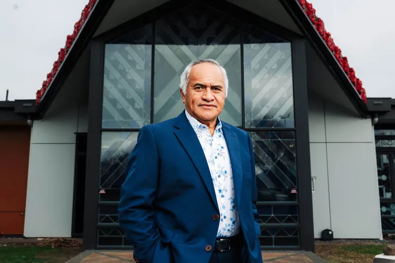 A man in a blue suit and floral shirt stands confidently in front of a modern building with geometric glass panels and red detailing along the roof.