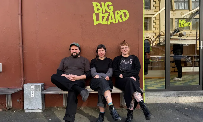 a man and two women sit on a wooden bench outside a shopfront that says "big lizard" in green writing