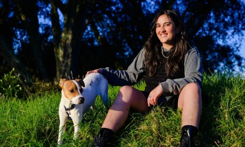A woman with long dark hair sits on grass, smiling at the camera, wearing a gray hoodie and black boots. She rests her hand on a white and brown dog standing beside her on the grassy hill, with trees in the background.