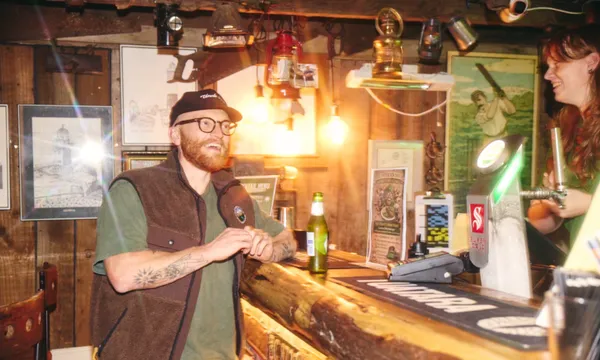 A man with glasses, a cap, and tattoos smiles while sitting at a rustic wooden bar, talking to a bartender. The bar is warmly lit with various decorations and framed pictures on the walls. A beer bottle is on the counter.