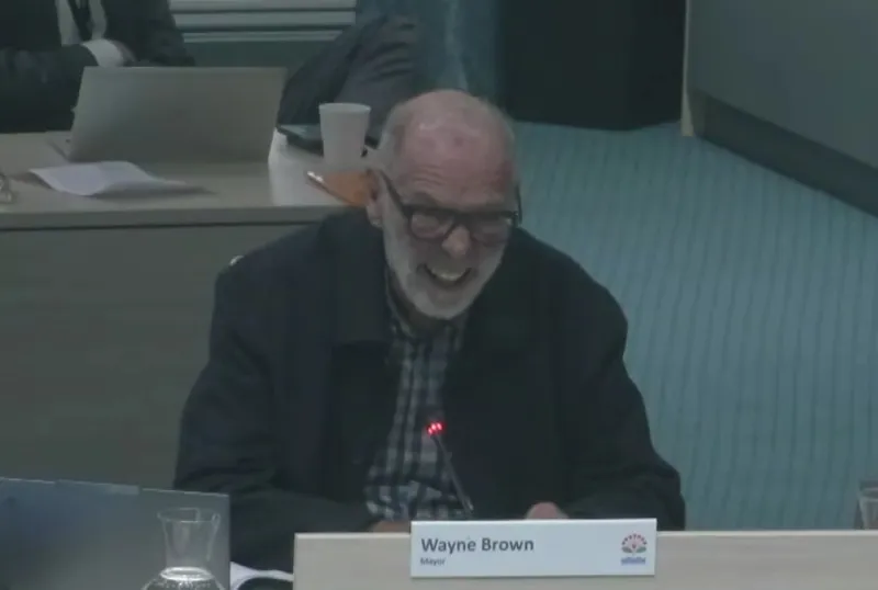 An older man with glasses and a beard, smiling while seated at a meeting table. A nameplate in front of him reads "Wayne Brown, Mayor." Papers and a glass of water are on the table.