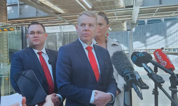 Three people in business attire stand together indoors, facing microphones from news outlets during a press conference. The central man in a suit and red tie appears to be speaking, with others standing attentively beside him.