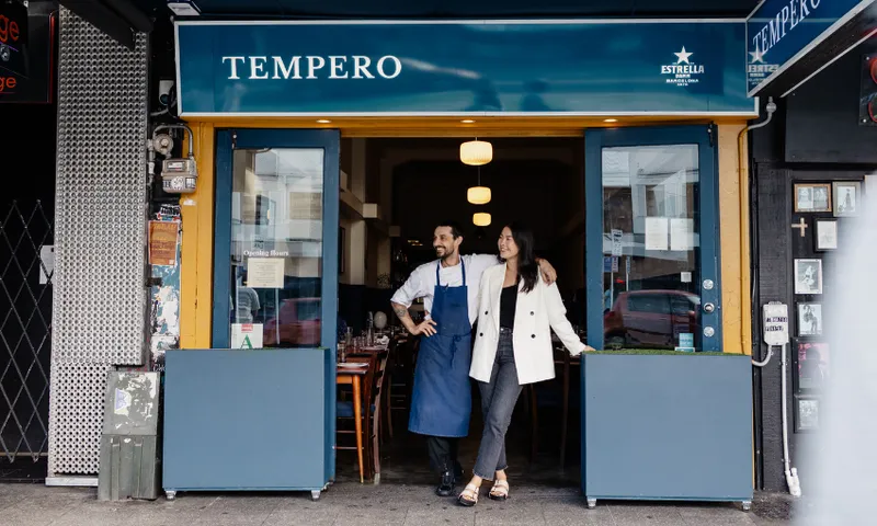 a man wearing an apron and a woman wearing a cream jacket and dark jeans stand in a restaurant doorway