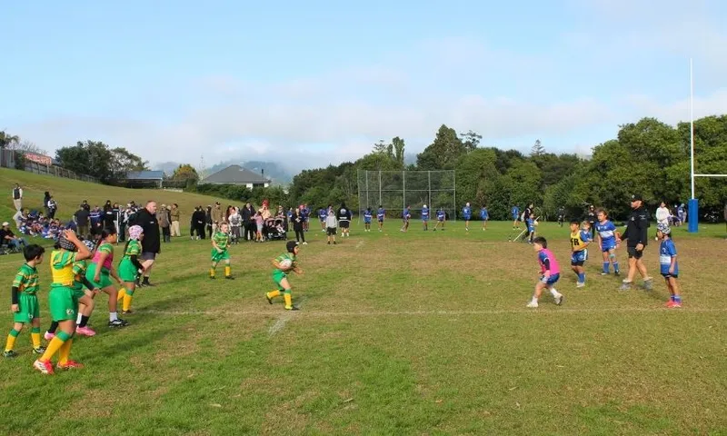 Children play a rugby league match on a grassy field, with one team in green and yellow and the other in blue and white. Spectators and coaches watch from the sidelines under a partly cloudy sky.