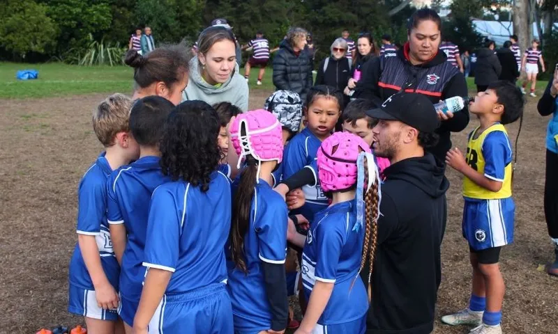 A youth rugby team in blue uniforms huddles together outdoors, listening to their coaches. Two children wear pink protective headgear. Adults and other children are seen in the background on the grassy field.
