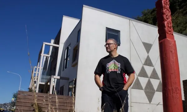 A person stands outside a modern white building with geometric designs, wearing glasses and a black T-shirt with a colorful design. A carved red post stands nearby under a clear blue sky.