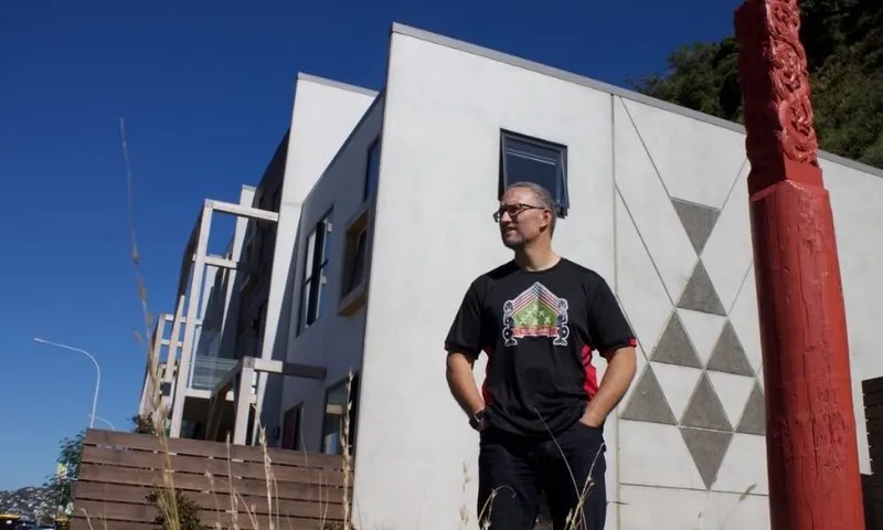 A person stands outside a modern white building with geometric designs, wearing glasses and a black T-shirt with a colorful design. A carved red post stands nearby under a clear blue sky.