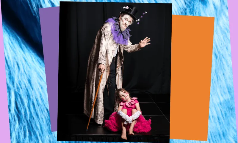 An elderly man in a top hat and gown appears on stage with his granddaugher, who is dressed as a pink flower, sitting at his feet