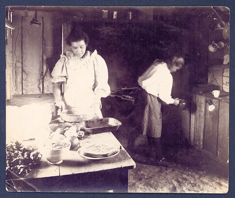 an old scan of a 1800s photograph showing a woman preparing food at a table with a woman in the background holding a jug of water