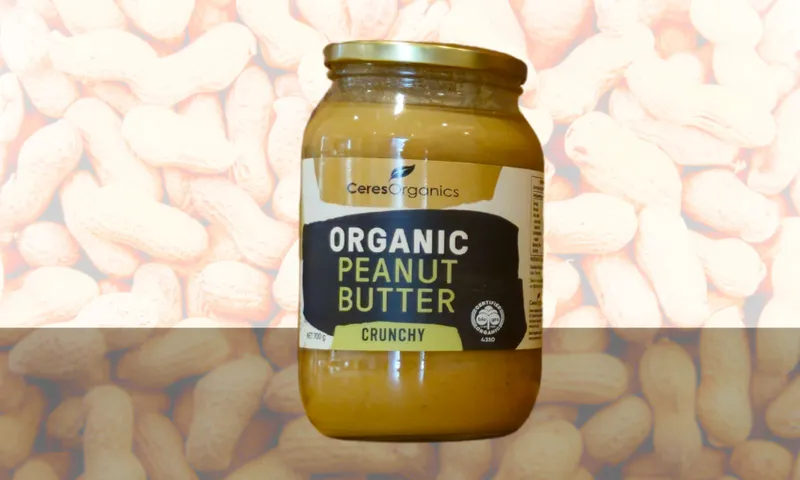 A glass jar of Ceres Organics Organic Peanut Butter Crunchy, with a label displaying the product name, is set against a background of whole peanuts in their shells.
