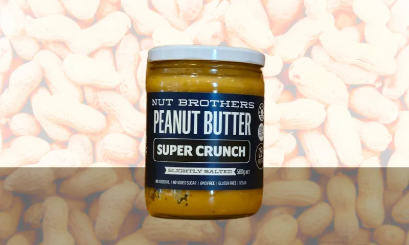A jar of Nut Brothers Peanut Butter Super Crunch, slightly salted, is displayed against a background of whole peanuts in their shells. The jar label is blue and white, and the jar contains 500 grams of peanut butter.