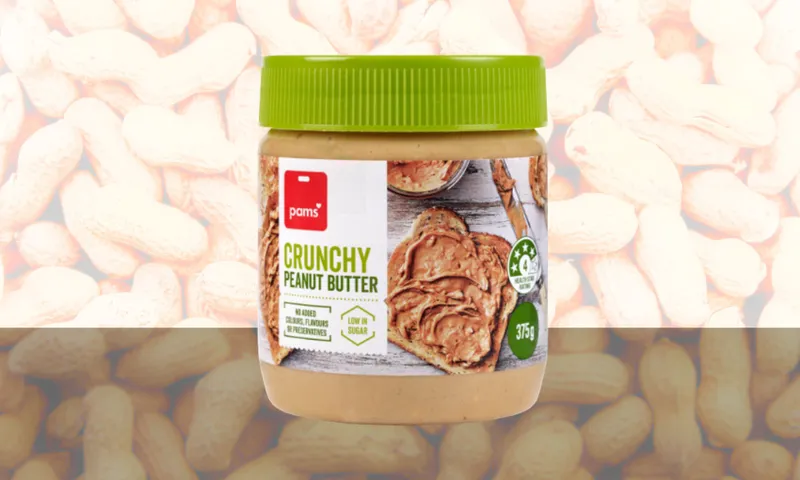 A jar of Pam’s Crunchy Peanut Butter with a green lid is placed in front of a peanut-patterned background. The label shows peanut butter spread on toast and emphasizes low sugar and sustainable sourcing.