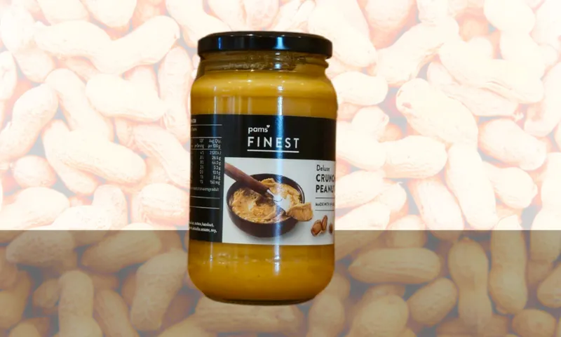 A jar of Pam's Finest Deluxe Crunchy Peanut Butter sits on a reflective surface, with a background featuring a pattern of whole peanuts.