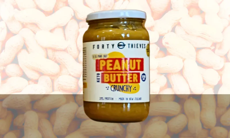 A jar of "Forty Thieves Crunchy Keto Peanut Butter" is centered against a blurred background of unshelled peanuts. The label highlights 29% protein and that it is made in New Zealand.