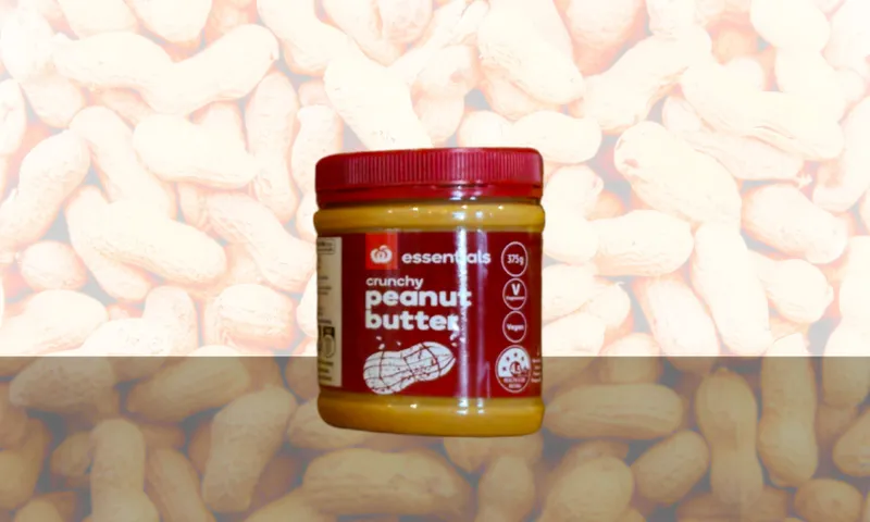 A jar of Essentials crunchy peanut butter with a red lid and label sits in front of a background filled with images of whole peanuts.
