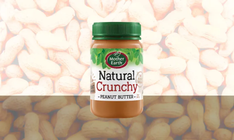 A jar of Mother Earth Natural Crunchy Peanut Butter with a green lid is shown against a background of unshelled peanuts. The label highlights that it contains no added sugar.