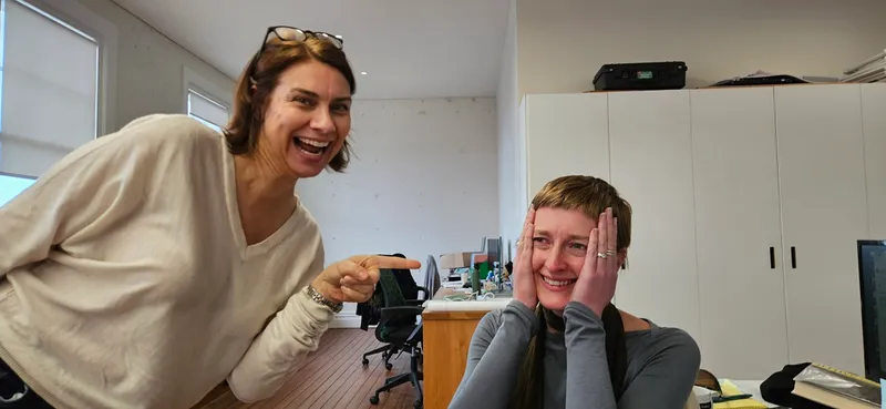 Two women in an office: one standing and smiling while pointing at the other, who is sitting at a desk, blushing and covering her cheeks with her hands, looking amused and surprised.