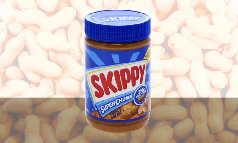 A jar of Skippy Super Chunk peanut butter with a blue lid is displayed in front of a background filled with unshelled peanuts.