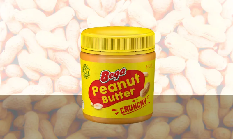 A yellow jar of Bega Crunchy Peanut Butter sits on a surface, with a peanut graphic on the label. The background shows a blurred image of many peanuts in their shells.