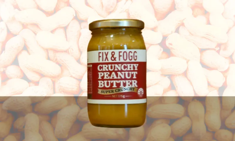 A jar of Fix & Fogg Crunchy Peanut Butter with a gold lid is displayed against a background filled with peanuts in their shells.