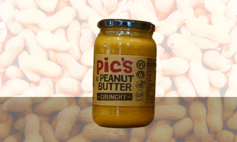 A jar of Pic's Crunchy Peanut Butter with a black lid is displayed against a background featuring images of peanuts in their shells. The label highlights its crunchy texture and gluten-free status.