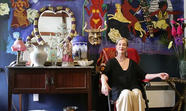 an older woman wearing a loose black top and cream skirt sits in an eclectic living room painted purple and filled with art on the walls