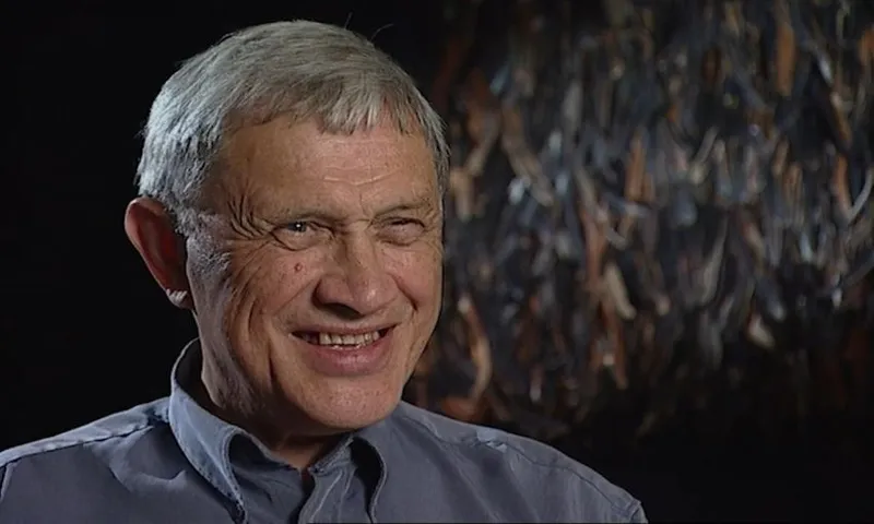 An older man with gray hair smiles warmly while wearing a gray collared shirt. The background is dark with abstract, textured patterns.