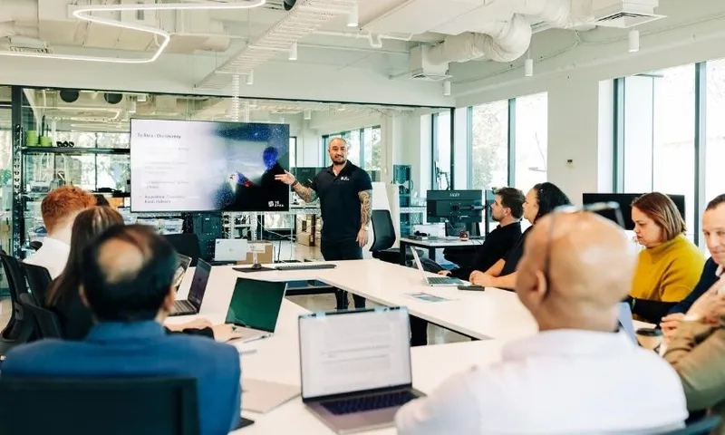 A man stands at the front of a modern conference room giving a presentation to a group of people seated around a table with laptops open, while a large screen displays a slide behind him.