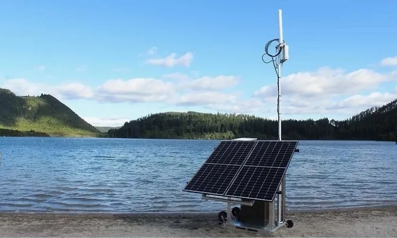 A portable solar panel system with an attached antenna stands on the sandy shore of a lake, surrounded by green hills and a partly cloudy blue sky.