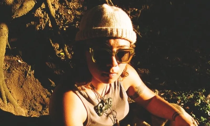 A person wearing a knit hat, sunglasses, and a sleeveless top sits outdoors in sunlight with shadows on their face. They have a large green pendant necklace and an arm tattoo.