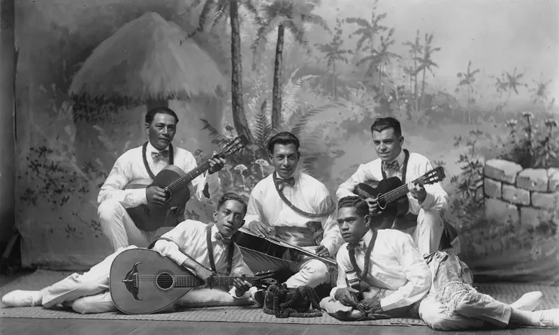 Five men dressed in white traditional clothing pose with string instruments in front of a painted tropical backdrop featuring palm trees, foliage, and a thatched hut.