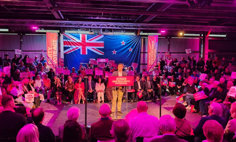 A speaker addresses a seated audience at an indoor event, with a large New Zealand flag displayed behind him. People hold signs, and pink lighting illuminates the scene. The main podium sign reads “UNLOCK NEW ZEALAND’S POTENTIAL.”.